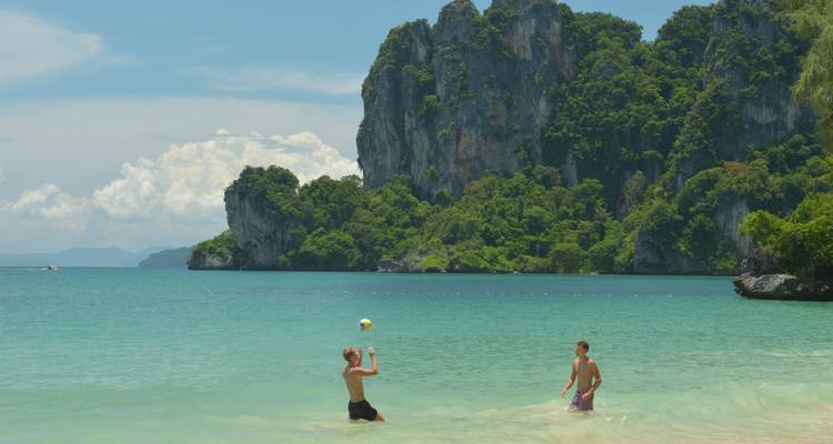 Two travelers play with a beach ball in clear turquoise water beneath limestone cliffs.