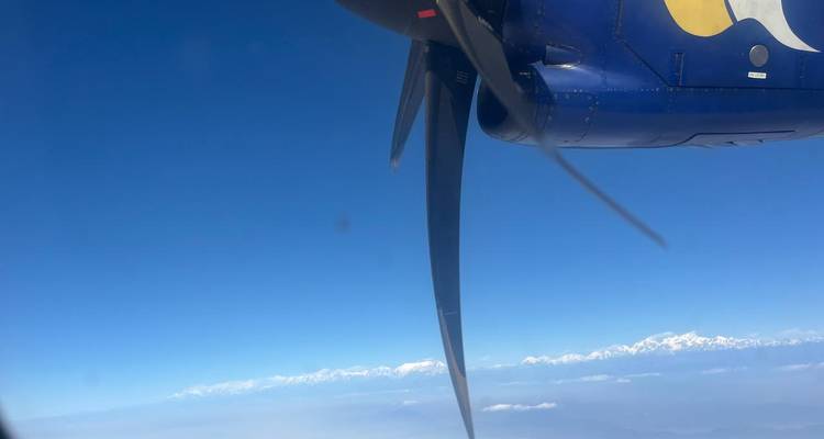 Aerial view from a propeller plane window showing the spinning blade against a clear blue sky and a distant snow-capped Himalayan range.