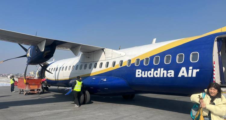 Passengers and ground crew beside a blue-and-yellow Buddha Air ATR aircraft parked on a sunny airport apron.