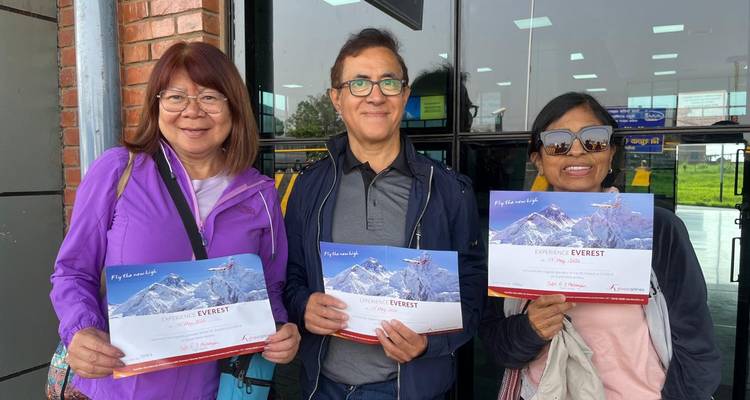 Three travellers proudly displaying Everest experience certificates outside an airport terminal entrance.