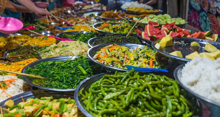 Farbenfrohe Schalen mit Gemüse, Obst und Currys, die auf einem belebten Nachtmarkt ausgestellt sind.