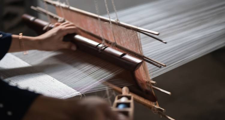 Nahaufnahme der Hände eines Webers, der an einem traditionellen Holzwebstuhl mit gespannten Seidenfäden arbeitet.