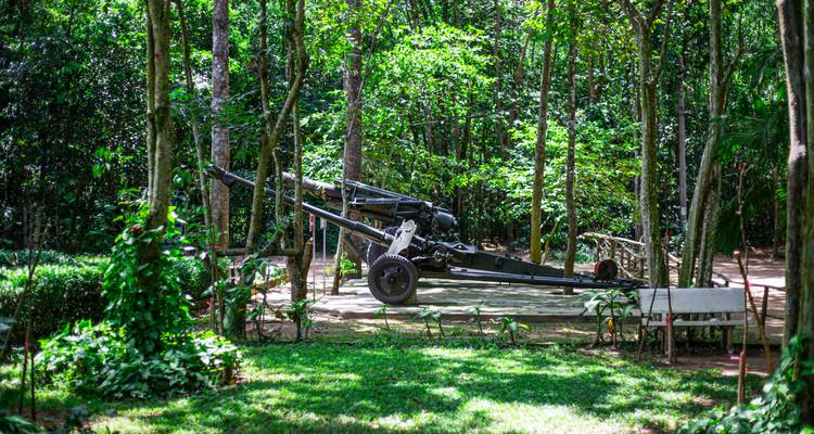 A preserved field artillery gun displayed on a concrete slab amid dense tropical forest greenery at an outdoor war site.