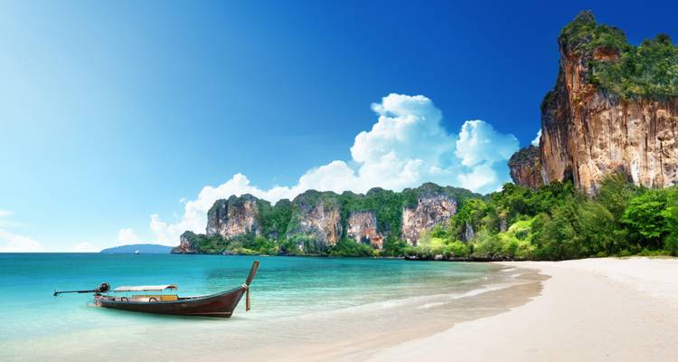 Serene tropical beach with long-tail boat anchored in turquoise water beneath limestone cliffs.
