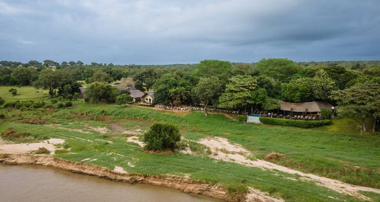 Plan de drone d'un complexe de lodge de safari en bordure de rivière entouré d'arbres denses sous un ciel nuageux.