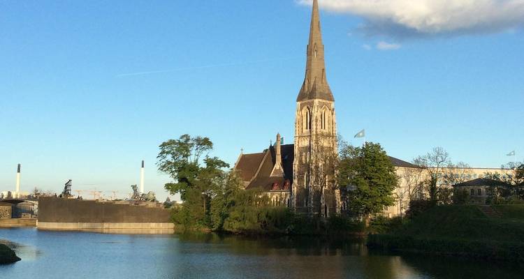 Una alta torre de iglesia de piedra y una característica de agua tipo foso se encuentran bajo un cielo de tarde despejado.