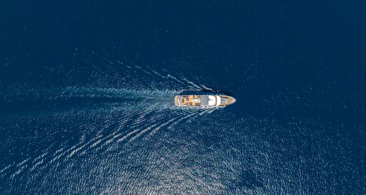 Vista aérea desde arriba de un yate navegando, dejando una estela en forma de V en el mar azul profundo.