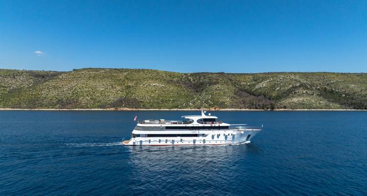 Perfil lateral de un yate de crucero frente a una costa montañosa verde bajo un cielo azul despejado.