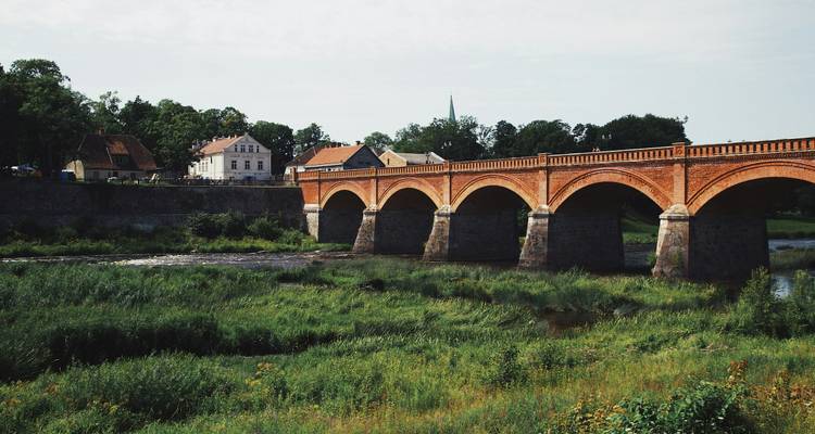 Historische Rotsteinfachwerkbrücke über ein grünes Flusstal mit Dorf im Hintergrund