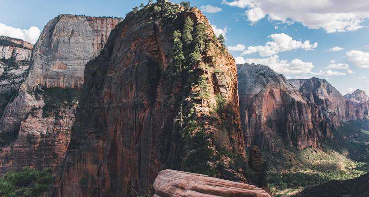 Des falaises de grès rouge imposantes et des canyons sous un ciel bleu partiellement nuageux avec une verdure ensoleillée parsemant les pentes.