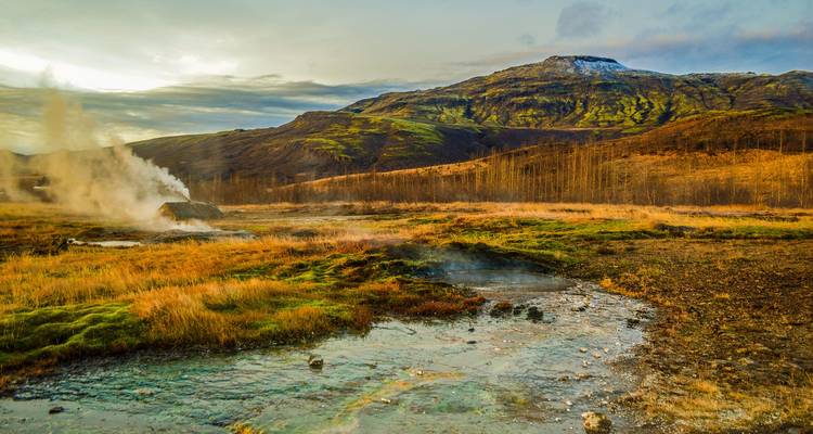 Steam billows from geothermal vent in colorful mossy valley beneath striped mountain