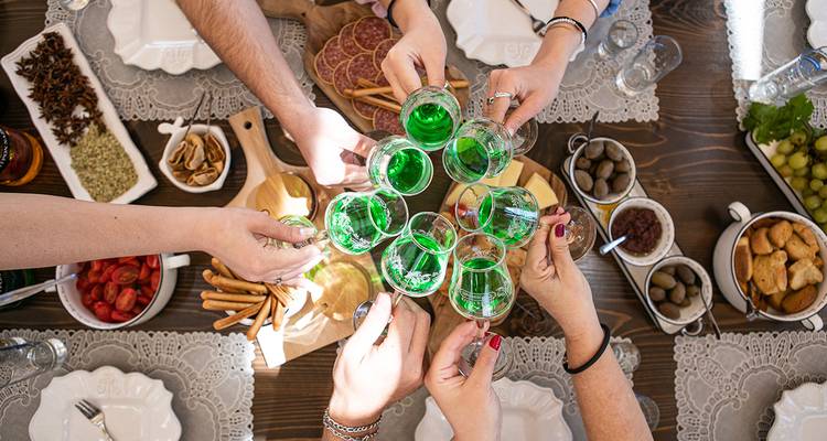 Vue de dessus de plusieurs personnes trinquant avec de petits verres d'une liqueur vert vif sur une table rustique remplie de collations méditerranéennes.