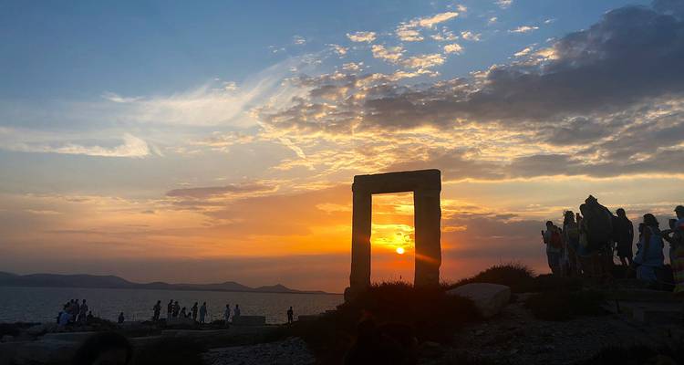 Les silhouettes de voyageurs se rassemblent près de l'ancienne Portara de pierre tandis que le soleil se couche sur la mer, peignant le ciel d'orange et de bleu.