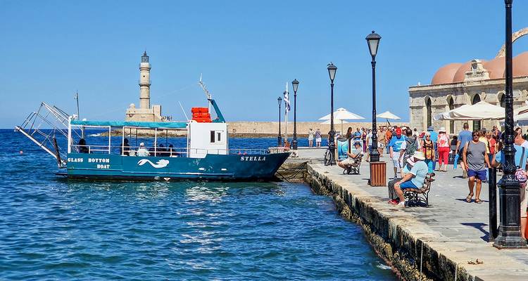 Une journée ensoleillée dans le vieux port vénitien de La Canée avec des touristes se promenant sur le quai en pierre près du phare et d'un bateau à fond transparent.