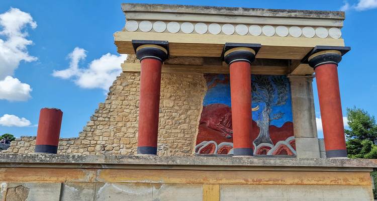 Les colonnes rouge vif et le mur aux fresques du palais reconstruit de Cnossos sous un ciel bleu parsemé de nuages épars.