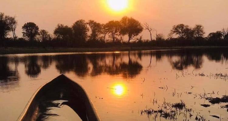 La proa de una canoa se desliza hacia una puesta de sol ardiente reflejada en aguas tranquilas bordeadas por árboles recortados contra el cielo.