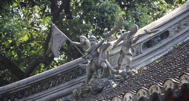 Stone carvings of ancient warriors poised atop a curved temple roof among trees.