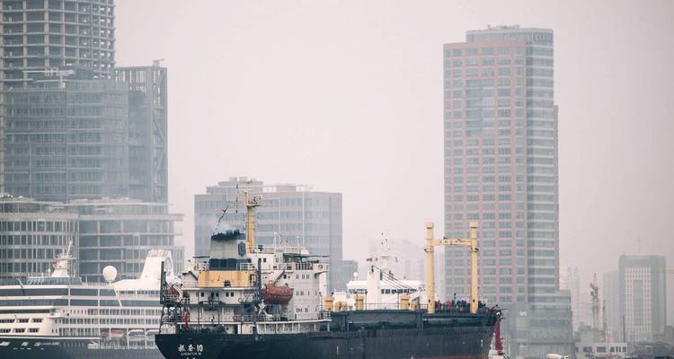 Cargo ship passes a hazy modern skyline along a broad river on an overcast day.