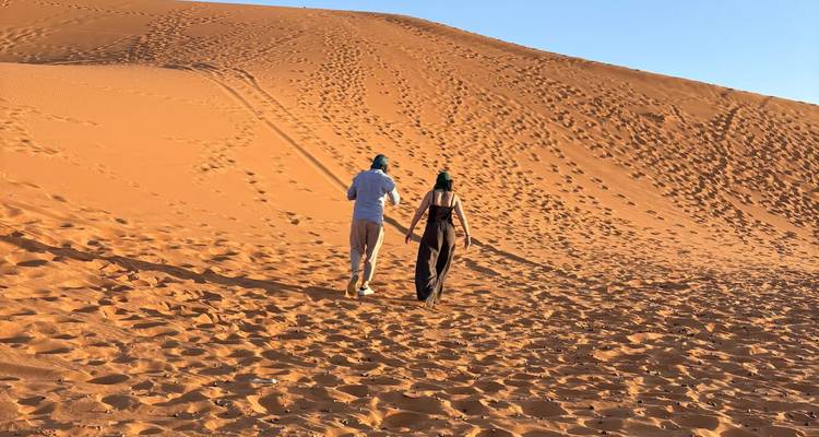 Pareja de la mano caminando por una duna de arena del Sahara iluminada por el sol y cubierta de huellas