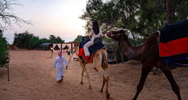 Un guide marchant à côté d'une femme montée sur un chameau en direction d'un campement désertique au crépuscule.