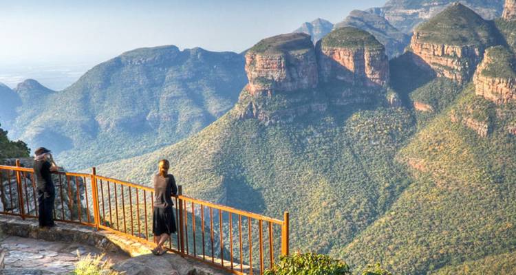 Dos visitantes están de pie en un mirador al borde de un acantilado con vistas a dramáticas montañas de cañón verde bajo una luz suave.