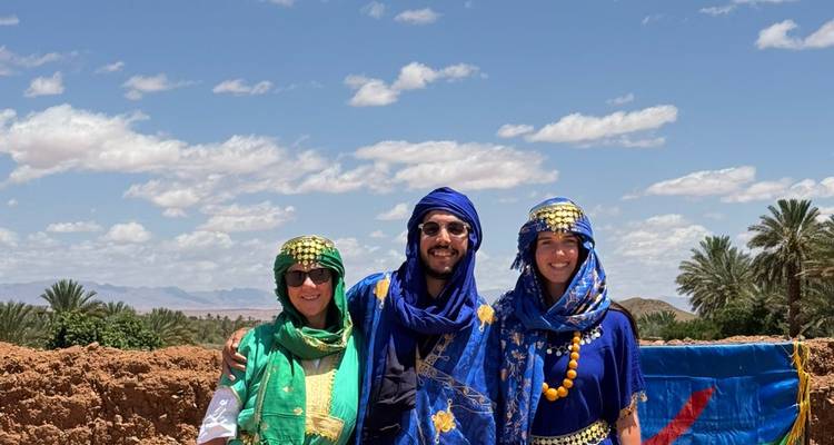 Group dressed in colorful Tuareg attire pose against desert oasis backdrop under bright sky