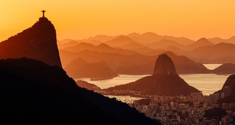 Dramatic sunset view over Rio de Janeiro with Christ the Redeemer silhouetted on Corcovado and Sugarloaf Mountain rising above the bay, city buildings in foreground