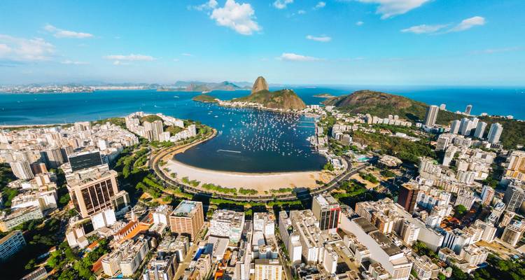 Aerial midday panorama of Rio de Janeiro showing Botafogo Bay, Sugarloaf Mountain, and surrounding cityscape under bright blue sky