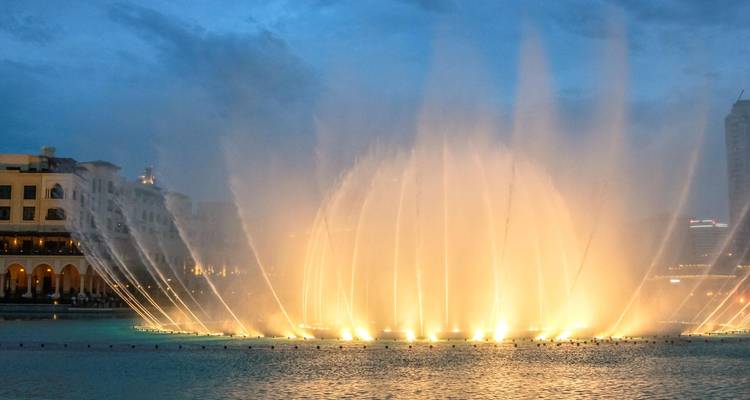 Beleuchtete Springbrunnen, deren Wasserstrahlen bei Einbruch der Dämmerung in die Luft schießen, neben Gebäuden am Wasser.