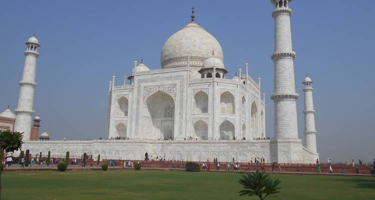 Seitliche Perspektive des Taj Mahal mit Besuchern, die entlang des Marmorsockels unter einem klaren blauen Himmel aufgereiht sind.