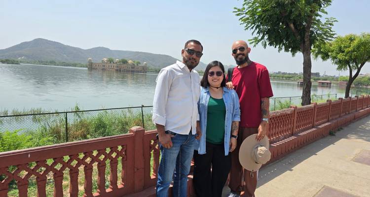 Three travellers smile for a photo beside a lakeside promenade with the Jal Mahal palace and hills behind.
