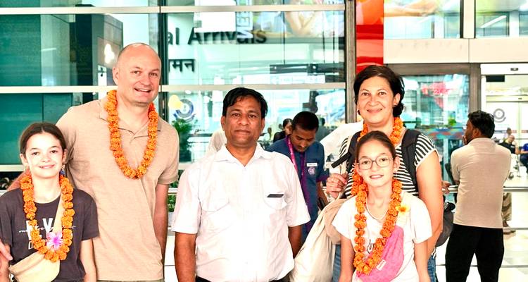 Family greeted with marigold garlands by their guide inside a brightly lit airport arrivals hall.