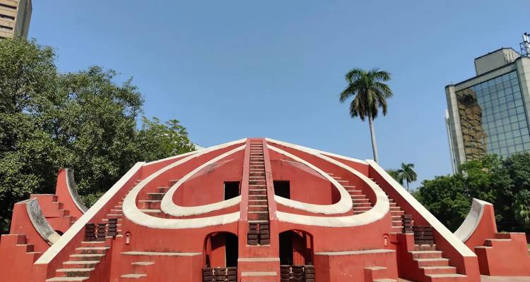 Red astronomical structure of Jantar Mantar framed by clear blue sky and palm trees.