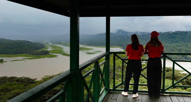 Dos mujeres con camisetas rojas disfrutan de una vista panorámica de la cuenca del río Chagres desde una plataforma de observación.