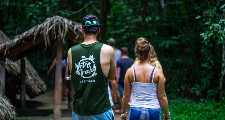 Back view of tourists walking through a forested war site, one wearing a Cu Chi travel tank top.