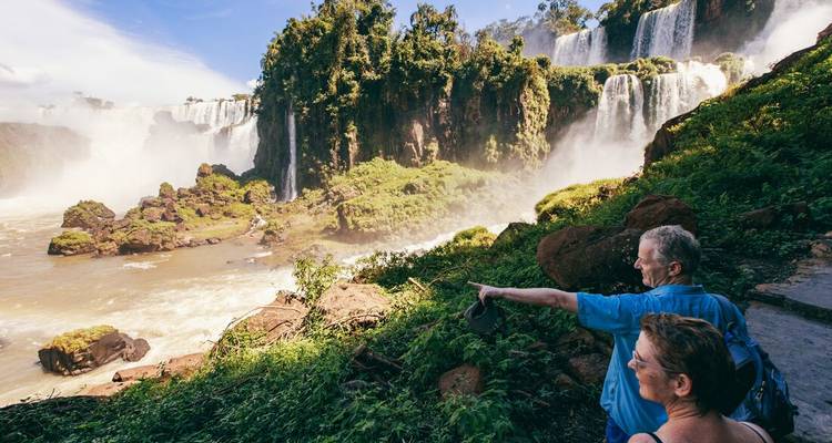 Travelling couple stands on lush trail admiring the misty power of Iguazu Falls.