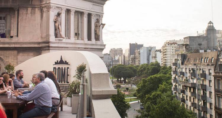 Groups of friends share drinks at a rooftop bar overlooking Buenos Aires cityscape at dusk.