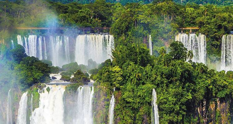 Wide view of multiple tiers of Iguazu Falls framed by verdant jungle and a faint rainbow.