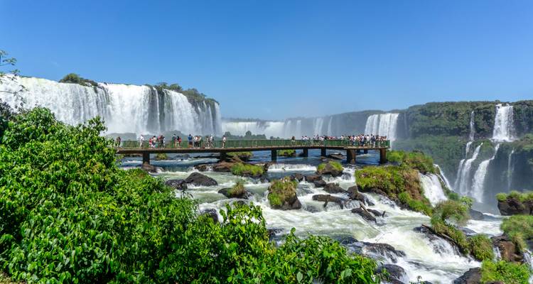 Tourists stand on a boardwalk surrounded by the thunderous cascades of Iguazu Falls under a blue sky.