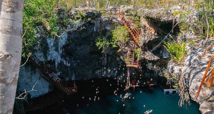 Diepe kalkstenen cenote met houten trappen die afdalen naar donkerblauw water en vogels die rondcirkelen.
