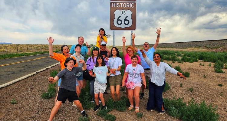 Blije toergroep springt onder het US 66 bord op een landelijk weggetje onder bewolkte hemel.