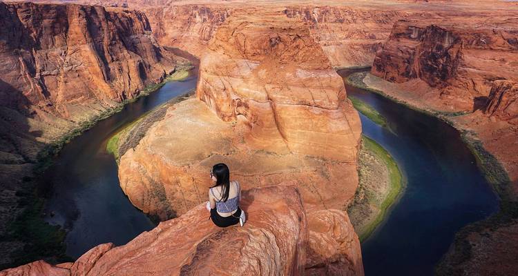 Bezoeker zit op de rand van een rode zandsteenrots boven de smaragdgroene bocht van Horseshoe Bend.
