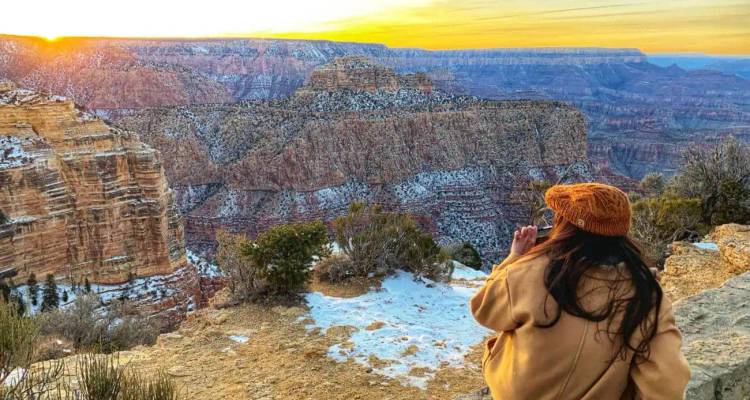 Traveller in orange beanie admires a snowy sunset panorama of the Grand Canyon from an exposed cliff edge.