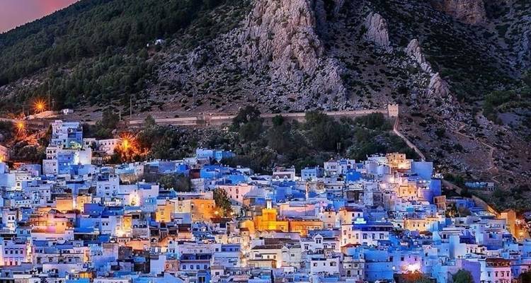 Avondaanzicht van de blauwe huizen van Chefchaouen die onder een rotsachtige berghelling zijn gegroepeerd, met warme stadslichten die glanzen.