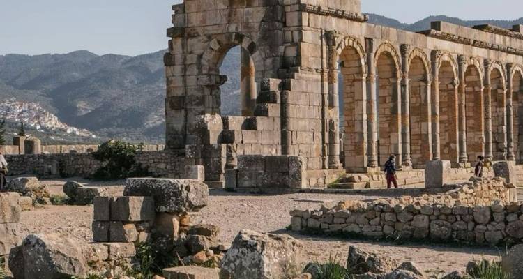 Bezoekers wandelen tussen de Romeinse ruïnes van Volubilis met stenen bogen en verspreid metselwerk in een landelijk landschap.