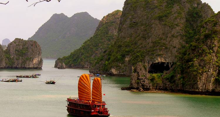 Traditionelles rotes Dschunkenboot mit orangefarbenen Segeln gleitet zwischen Kalksteinfelsen in der Halong-Bucht.