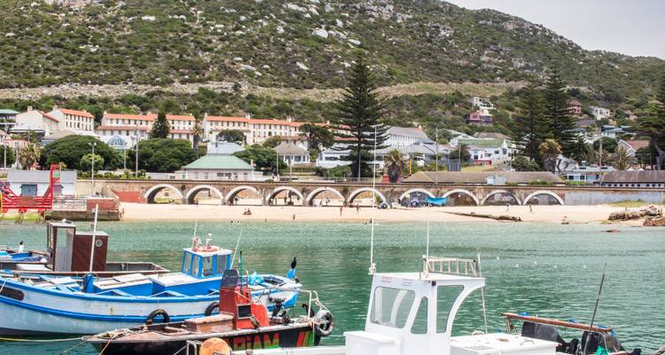 Vissersboten afgemeerd in een rustige haven met bergen op de achtergrond en een zandstrand in een kustdorpje.