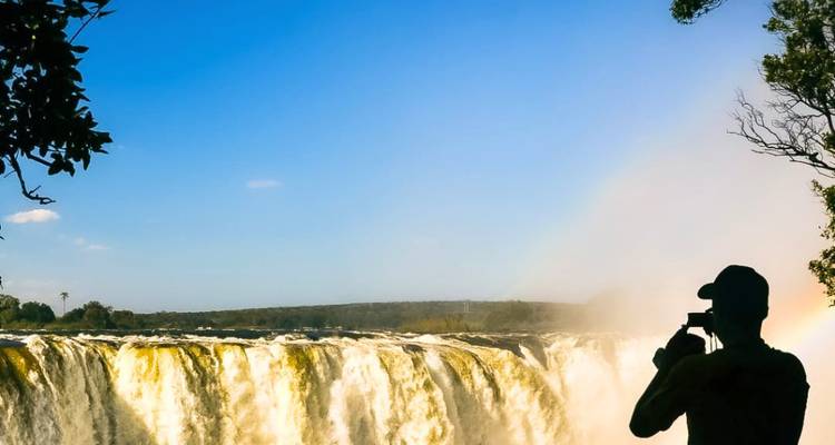 Silhouet van een fotograaf die het brullende gordijn van de Victoria Falls vastlegt onder een strakblauwe hemel.