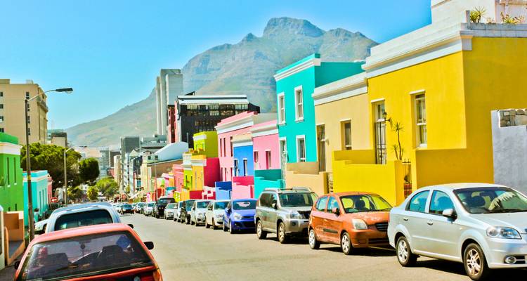 Felgekleurde huisjes langs de Bo-Kaap straat met bergen op de achtergrond en geparkeerde auto's.