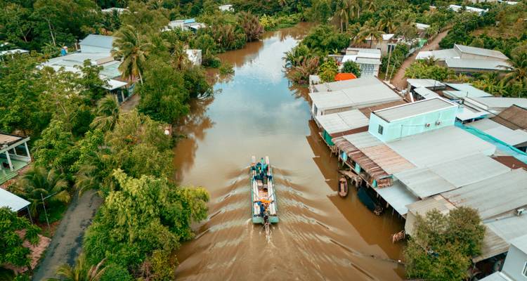Aerial view of a long-tail boat cruising down a muddy canal lined with tropical houses and lush palms in the Mekong Delta.